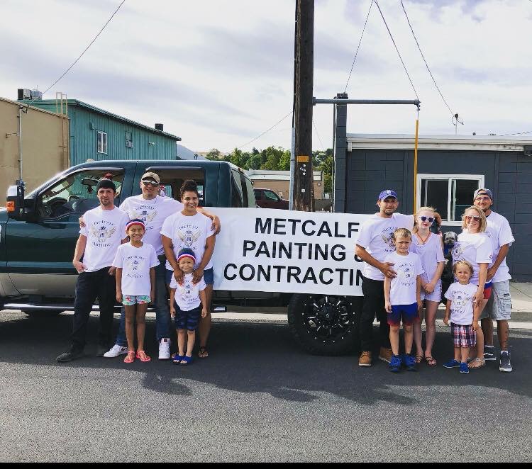 The Metcalf crew and family in matching shirts holding a Metcalf Painting and Contracting banner beside their truck